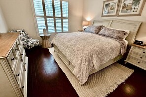 Un dormitorio con cama, mesitas de noche, cómoda, ventana con persianas e imágenes enmarcadas en la pared.