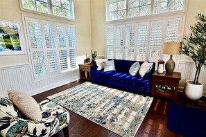 Un salon avec un canapé bleu, un tapis à motifs et une grande fenêtre avec des volets.