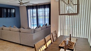 A living room with a sectional sofa, a dining table with chairs, a ceiling fan, and a view through the window.