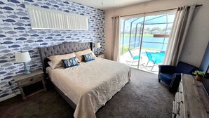 A bedroom with a large bed, a nightstand, a lamp, a blue chair, and a view of a pool through large windows.