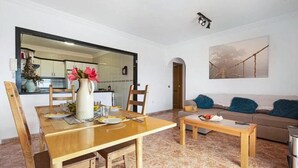 Ein Wohnzimmer mit Holzesstisch, einer Couch und einem Couchtisch. Durch einen offenen Durchgang ist eine Küche erkennbar.