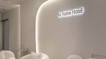A modern hotel lobby with a curved wall displaying the name 'At Home Hotel'. There are plush white sofas and a round coffee table.