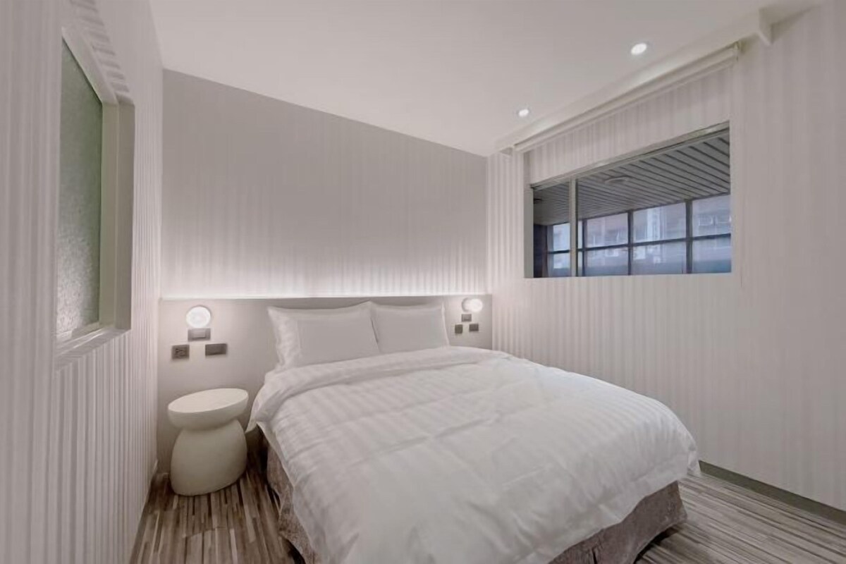 A modern hotel room with a large bed, a round bedside table, a window with blinds, and a small round toilet.