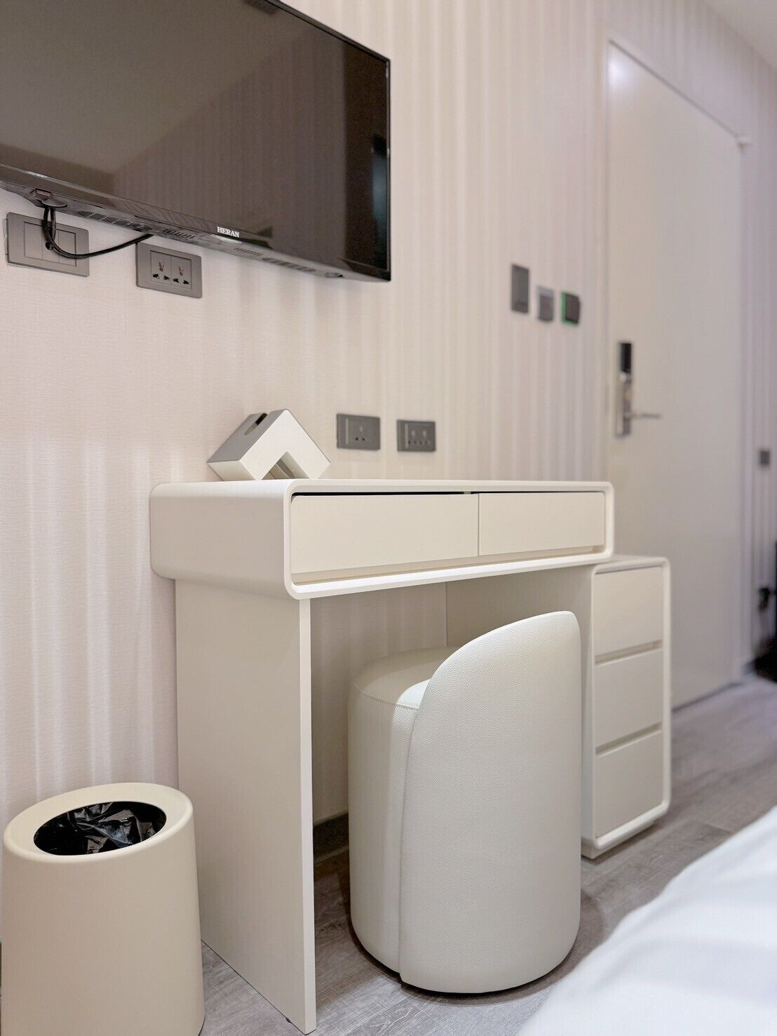 Ein modernes Hotelzimmer mit einem weißen Schreibtisch, einem Stuhl, einer Mülltonne, einem Spiegel und einem Fernseher, der über dem Schreibtisch angebracht ist.