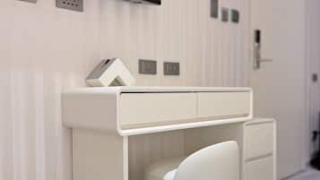 Ein modernes Hotelzimmer mit einem weißen Schreibtisch, einem Stuhl, einer Mülltonne, einem Spiegel und einem Fernseher, der über dem Schreibtisch angebracht ist.