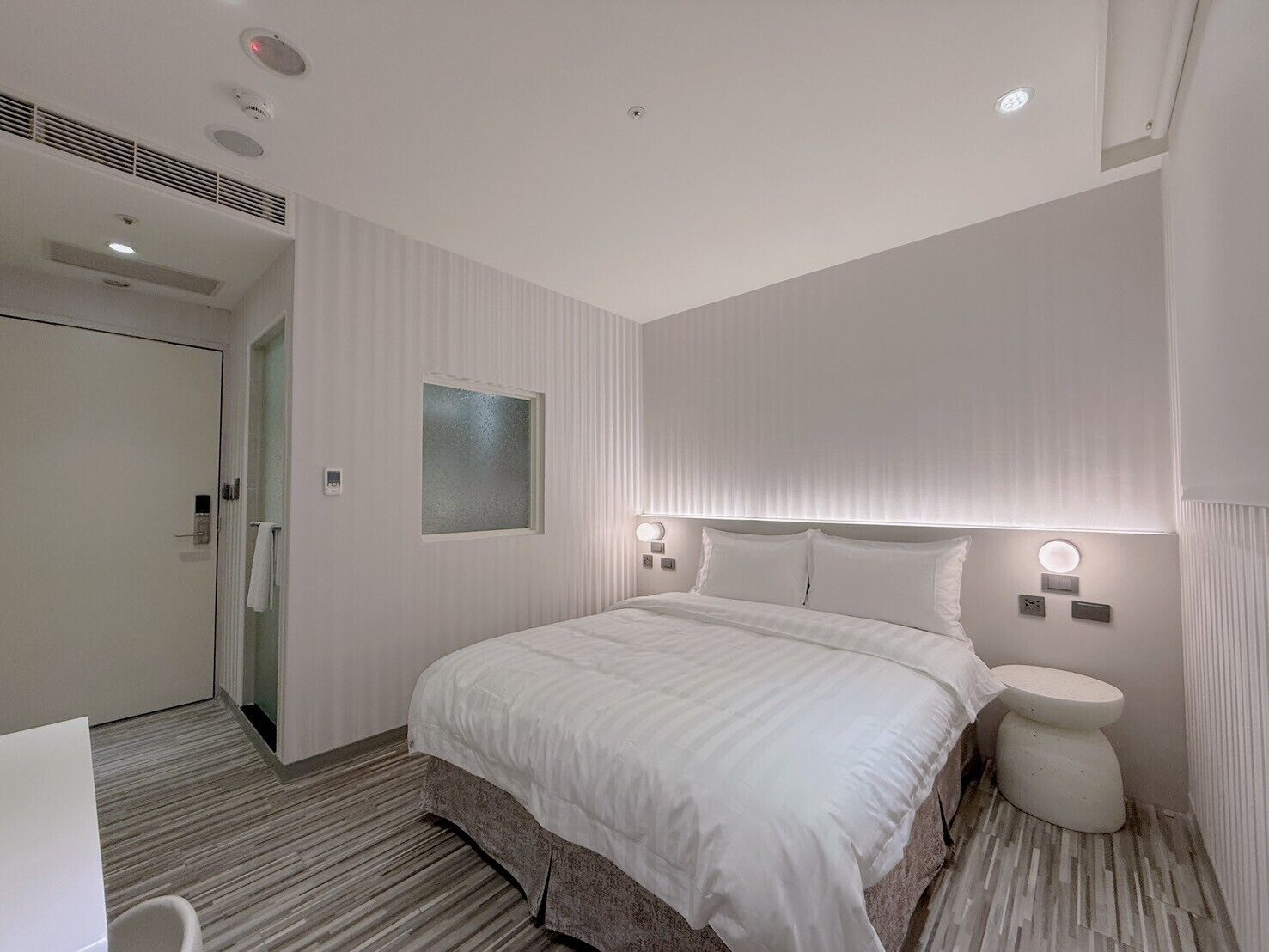 A modern hotel room with a large bed, a bedside table, a small bedside lamp, and a toilet.