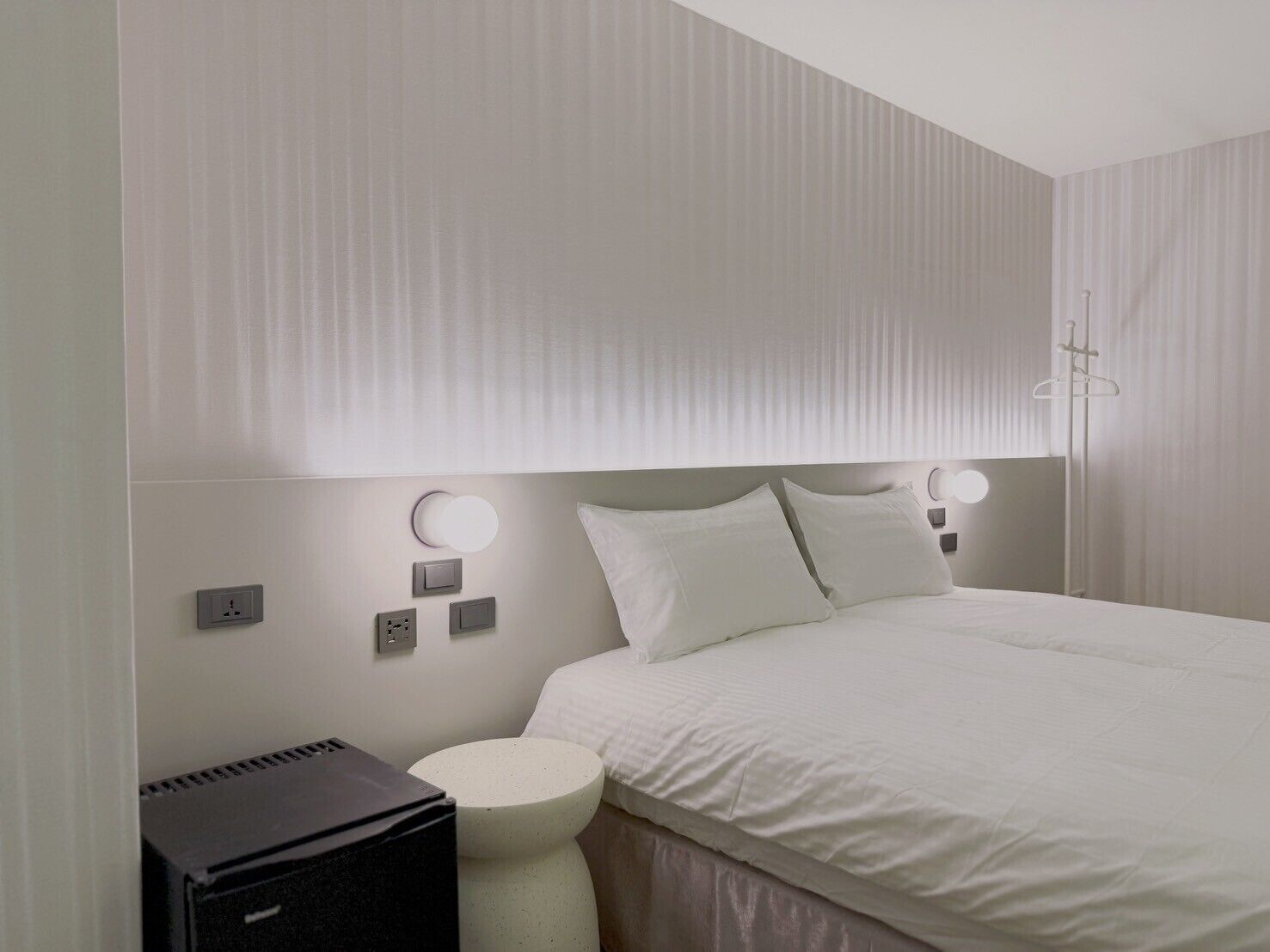 A modern hotel room with a large bed, bedside lamps, a round bedside table, and a black suitcase.