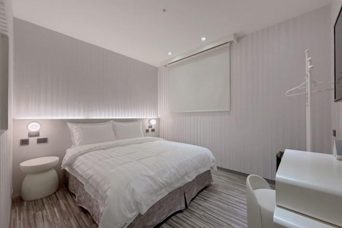 A modern hotel room with a large bed, bedside tables, a white wardrobe, and a large window.