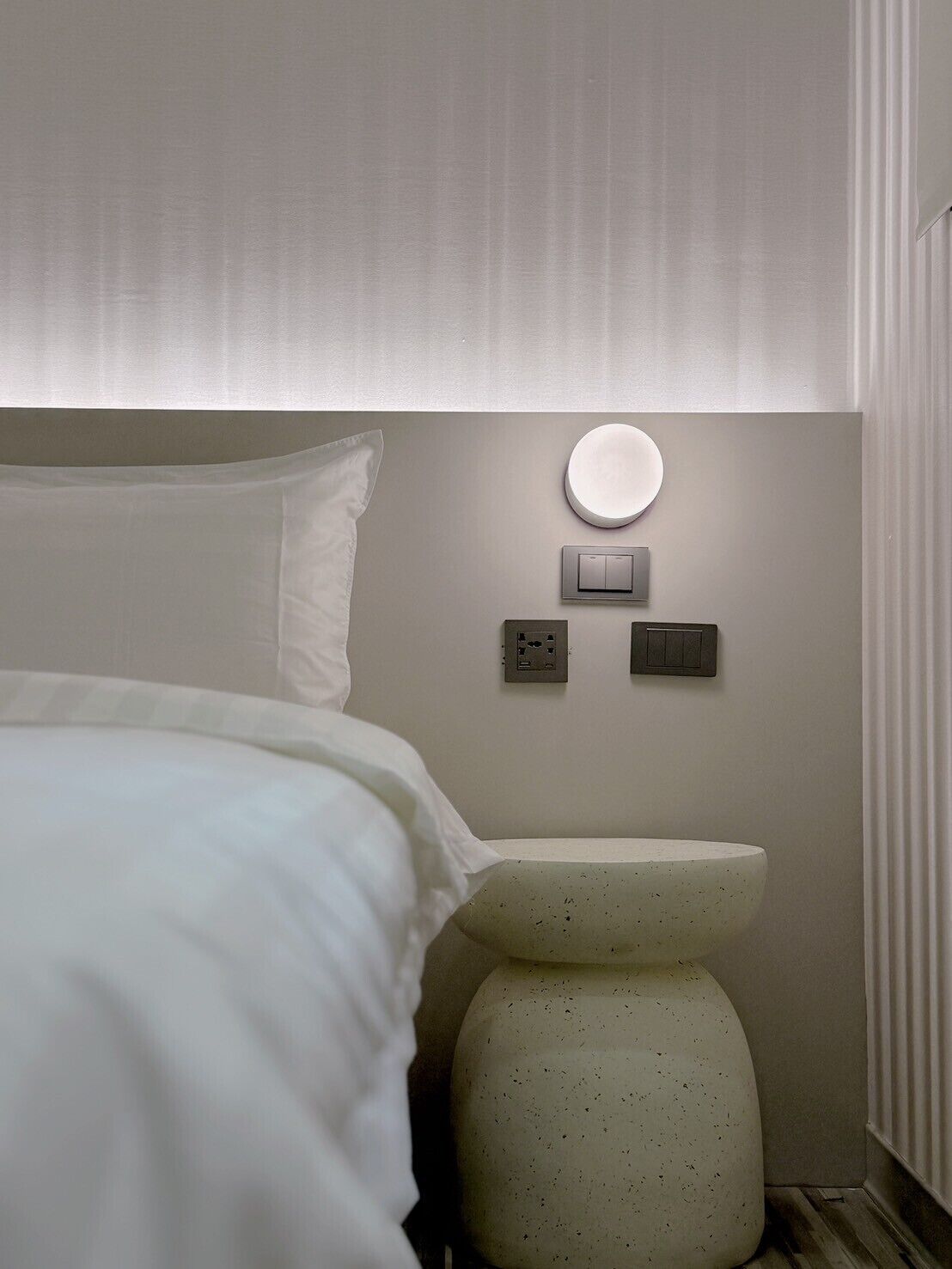 Moderni hotellihuone, jossa on sänky, pyöreä seinään kiinnitetty valaisin, termostaatti ja wc.