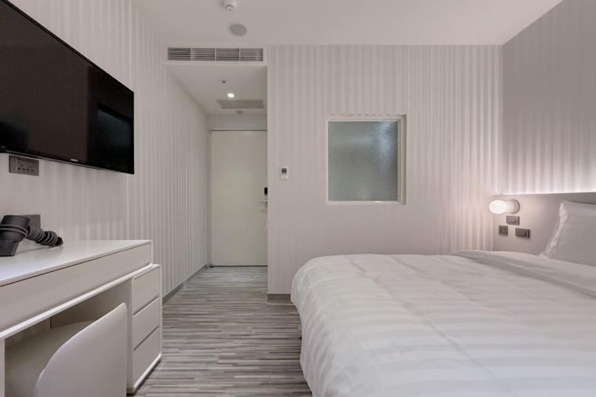 A modern hotel room with a bed, a desk with a chair, a flat-screen TV mounted on the wall, and a bedside table with a lamp.