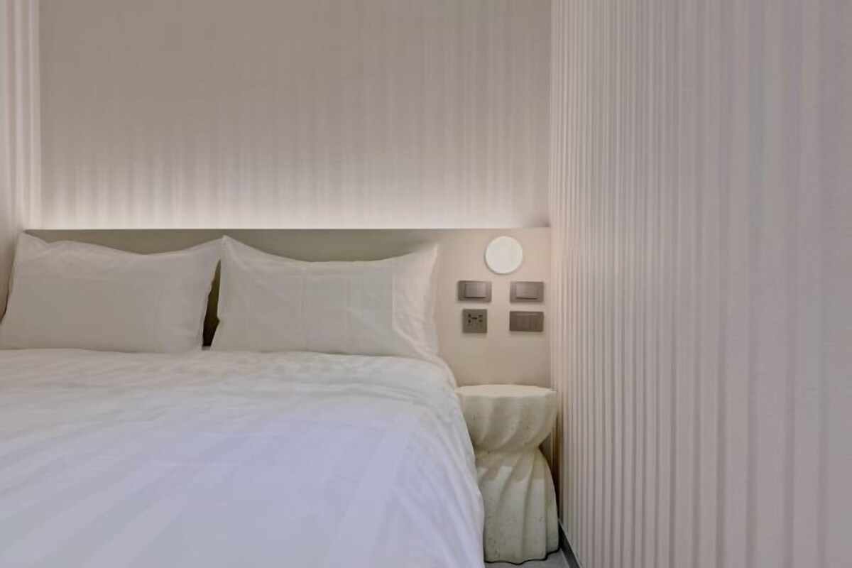 A neatly made bed with white linens, a round wall clock, and a small bedside table with a lamp.