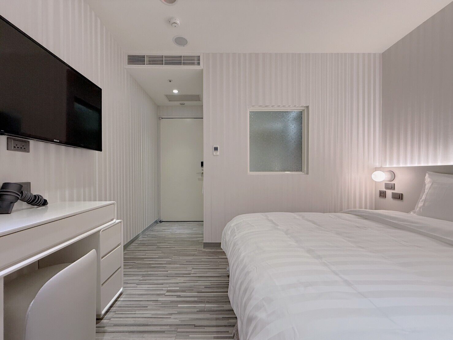 Een moderne hotelkamer met een flatscreen televisie, een bureau met lamp, een bed met hoofdeinde en een raam met gordijnen.