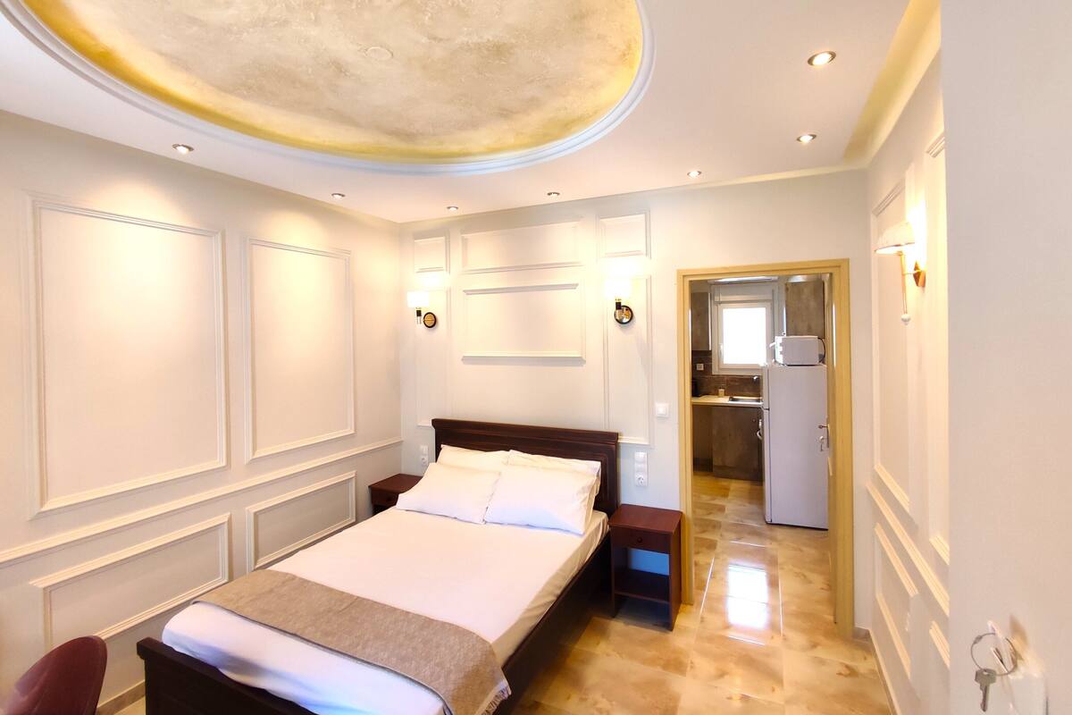 A hotel room with a bed, bedside tables, a nightstand, a chair, a bathroom with a sink and mirror, and a kitchen area.
