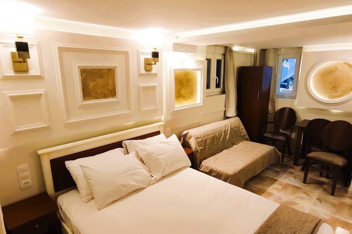 A hotel room with a large bed, a small table, and a chair. There are wall sconces and a window with curtains.