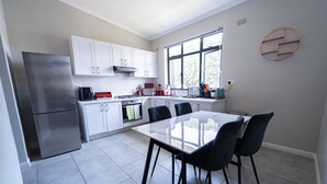 Una cocina moderna con armarios blancos, un refrigerador de acero inoxidable, una mesa de comedor de vidrio con cuatro sillas y una ventana con persianas.