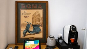 Ein gerahmtes Bild des Kolosseums mit dem Text 'Roma Vacanze 1958' und einer Illustration eines Vintage-Rollers. Auf einem Schreibtisch stehen ein Tablet, eine Kaffeemaschine und eine Fernbedienung.
