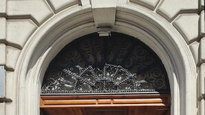 Ein rundbogiger Durchgang mit einem verzierten Metallwappen, Holztüren und einem gefliesten Boden.