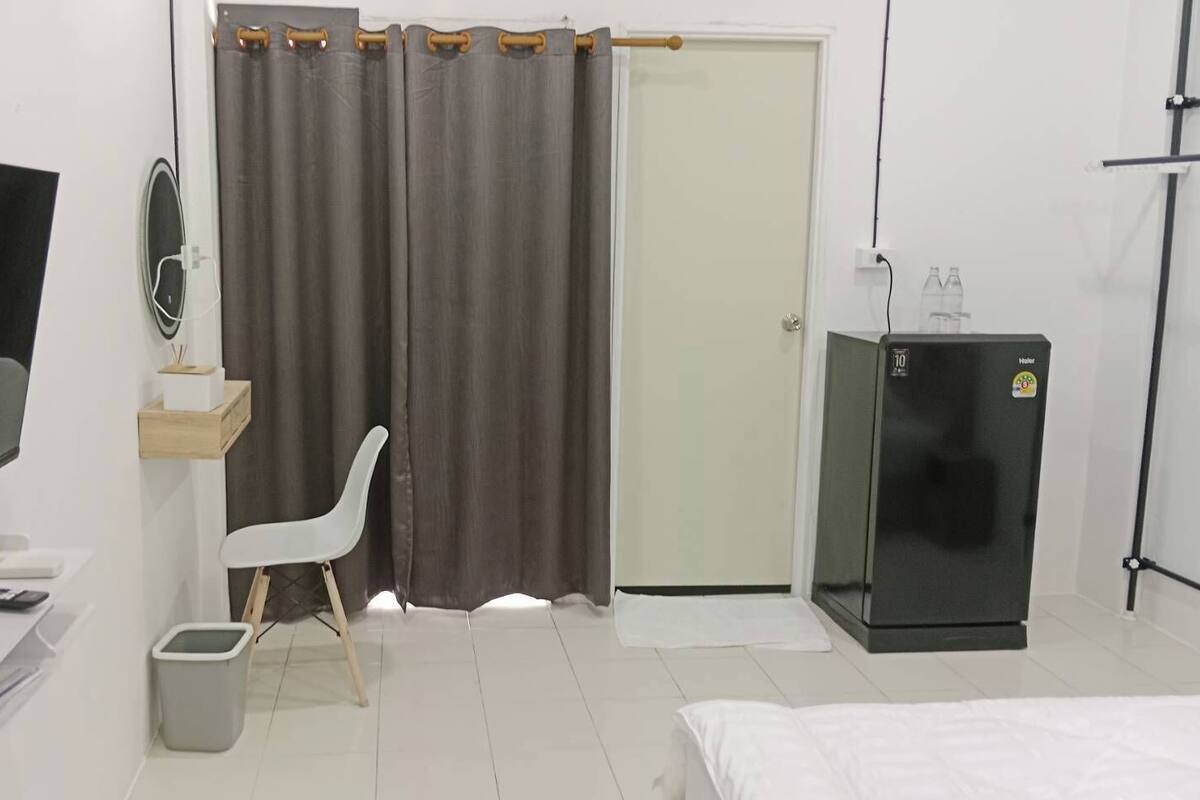 Habitación de hotel con cama, escritorio con silla, televisión, refrigerador, cortina y puerta.