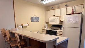 Une cuisine de taille réduite avec un îlot central, des meubles blancs, un four à micro-ondes, un réfrigérateur et un panneau affiché sur le mur.