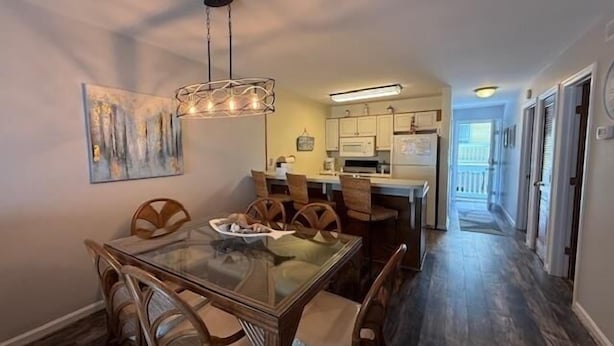 Un coin repas avec une table en verre et des chaises en bois, une cuisine à l’arrière-plan et un couloir menant à une porte.