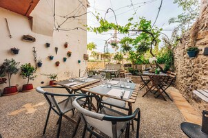 Un patio al aire libre con mesas y sillas de madera, rodeado de plantas en macetas y un muro de piedra.