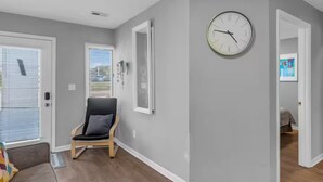 Un salon moderne avec un mur gris, un plancher en bois, un fauteuil noir, une horloge et une porte donnant sur une autre pièce.