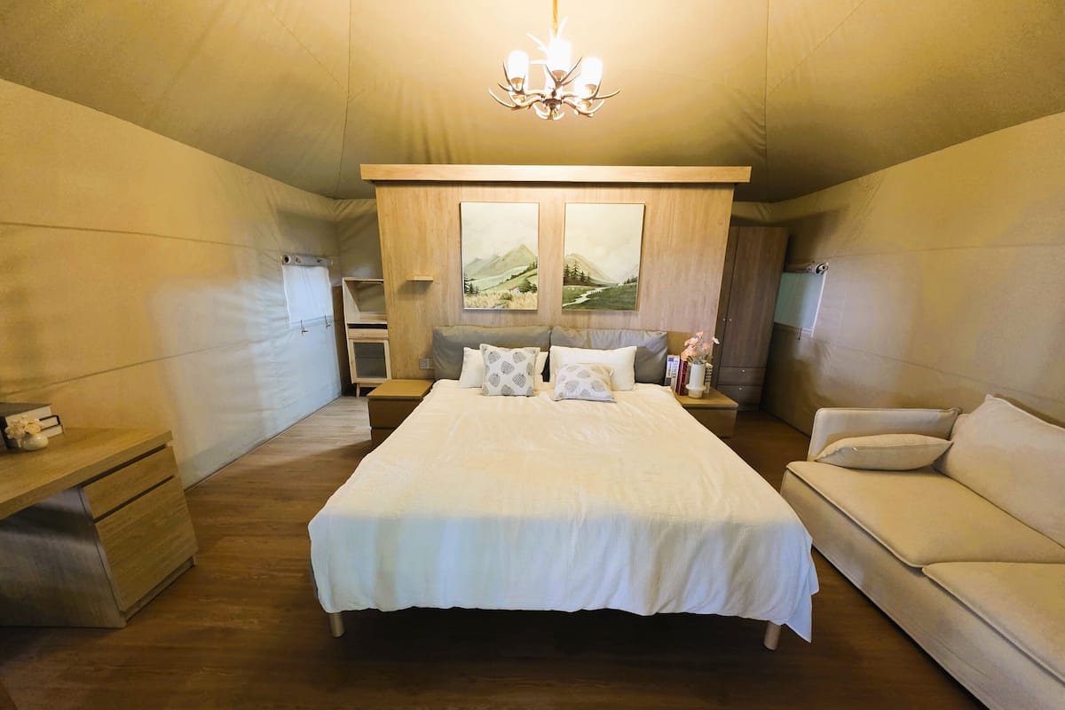 A bedroom with a large bed, a wooden nightstand, a bedside table, a sofa, and a window with a view of the outdoors.