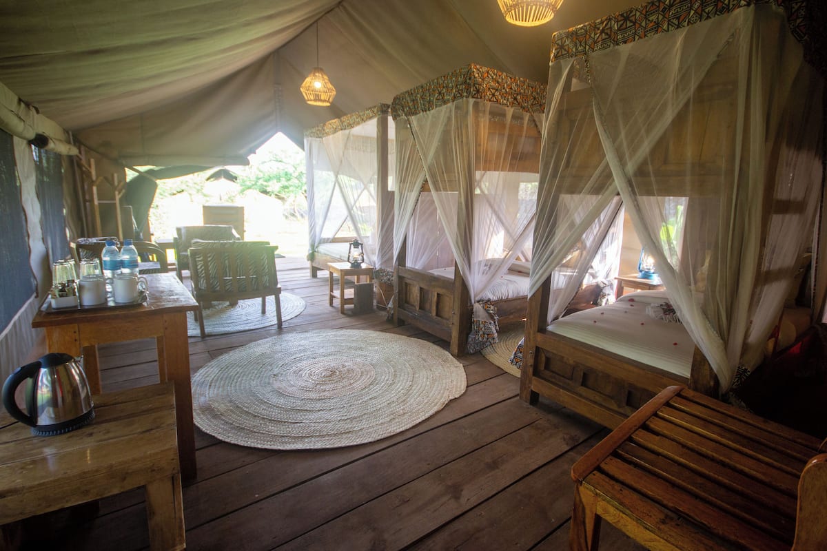 A room with a wooden floor, a round woven rug, a bed with a canopy, a wooden bench, and a table with a kettle and cups.
