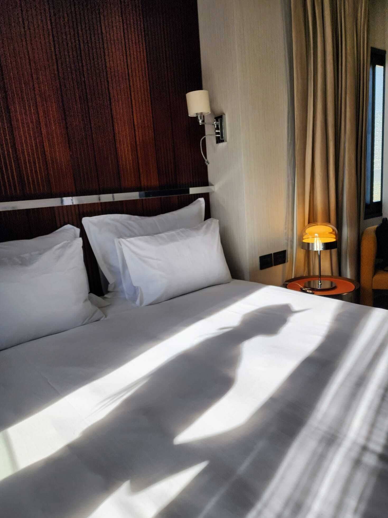 A hotel room with a large bed, two pillows, a nightstand with a lamp, and a wooden headboard.