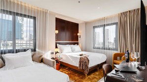 A hotel room with two beds, a desk, a chair, a small table with a bottle and glass, and a view of the city through large windows. - Fiori 41 (Tel Aviv)