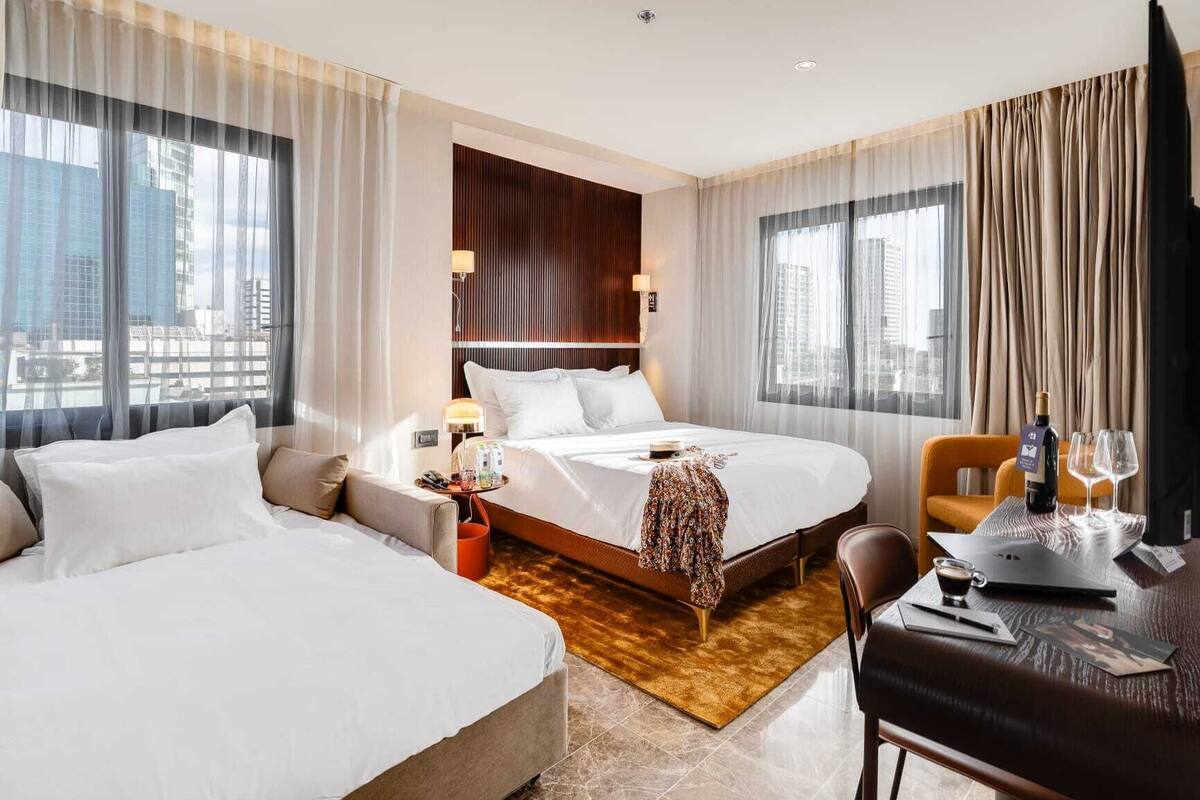 A hotel room with two beds, a desk, a chair, a small table with a bottle and glass, and a view of the city through large windows.