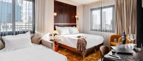 A hotel room with two beds, a desk, a chair, a small table with a bottle and glass, and a view of the city through large windows.