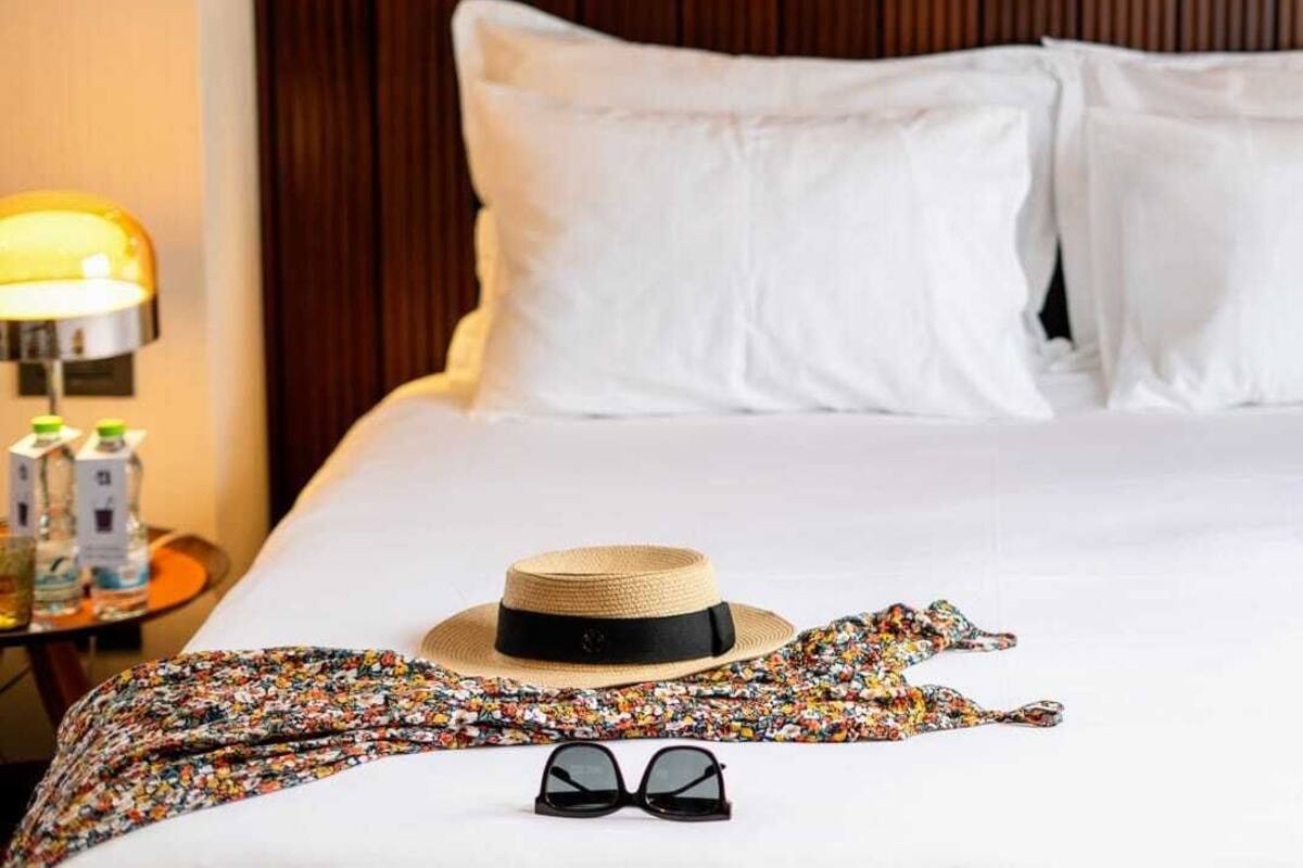 A neatly made bed with white linens, a straw hat, a colorful scarf, and black sunglasses on the bed.