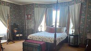 En seng med himmelseng, et blomstret sengetæppe, en træbænk, et lille bord og en lampe.