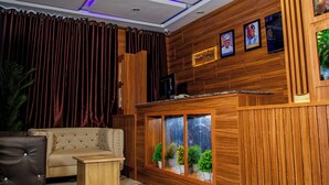 Ein Zimmer mit einem hölzernen Empfangsschreibtisch, einer gepolsterten Couch, einem kleinen Tisch und einem großen Fenster mit Topfpflanzen.