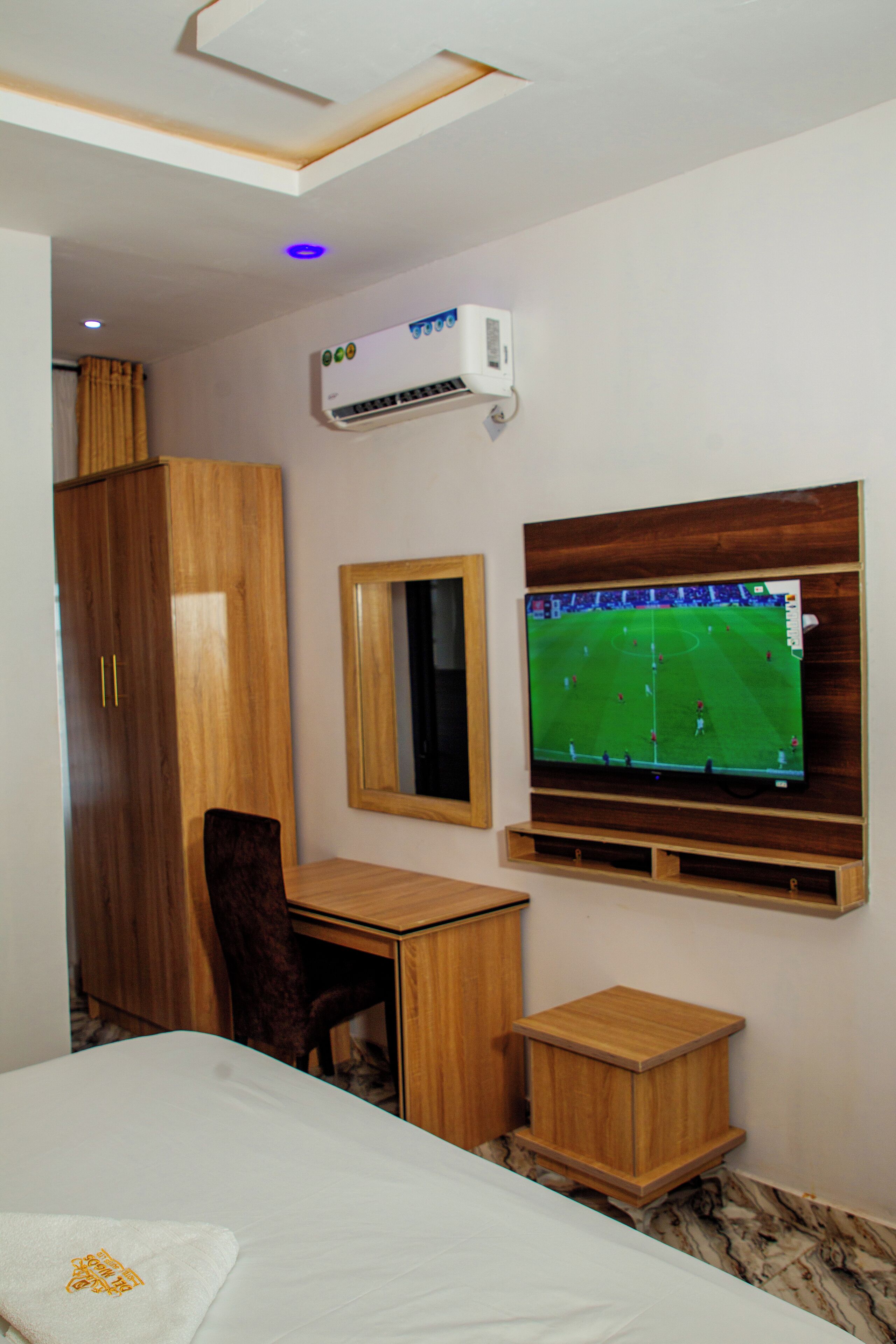 A hotel room with a bed, a wooden desk, a television mounted on the wall, a wardrobe, and an air conditioning unit.