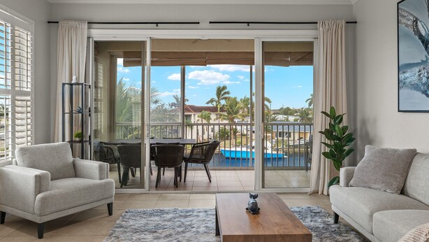 Ein modernes Wohnzimmer mit Sofa, Sessel und Couchtisch, ein Balkon mit Blick auf einen Pool und ein Essbereich mit Stühlen und Tisch.