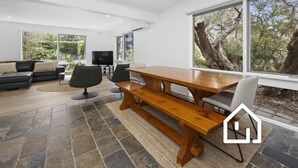 A modern living room with a wooden dining table, black leather sofa, and a large window offering a view of trees. - Albany Beachside Retreat *1 NIGHT FREE* (Sorrento)