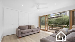 A modern living room with a grey sofa, a wooden floor, and a sliding glass door leading to an outdoor patio with a table and umbrella. - Albany Beachside Retreat *1 NIGHT FREE* (Sorrento)
