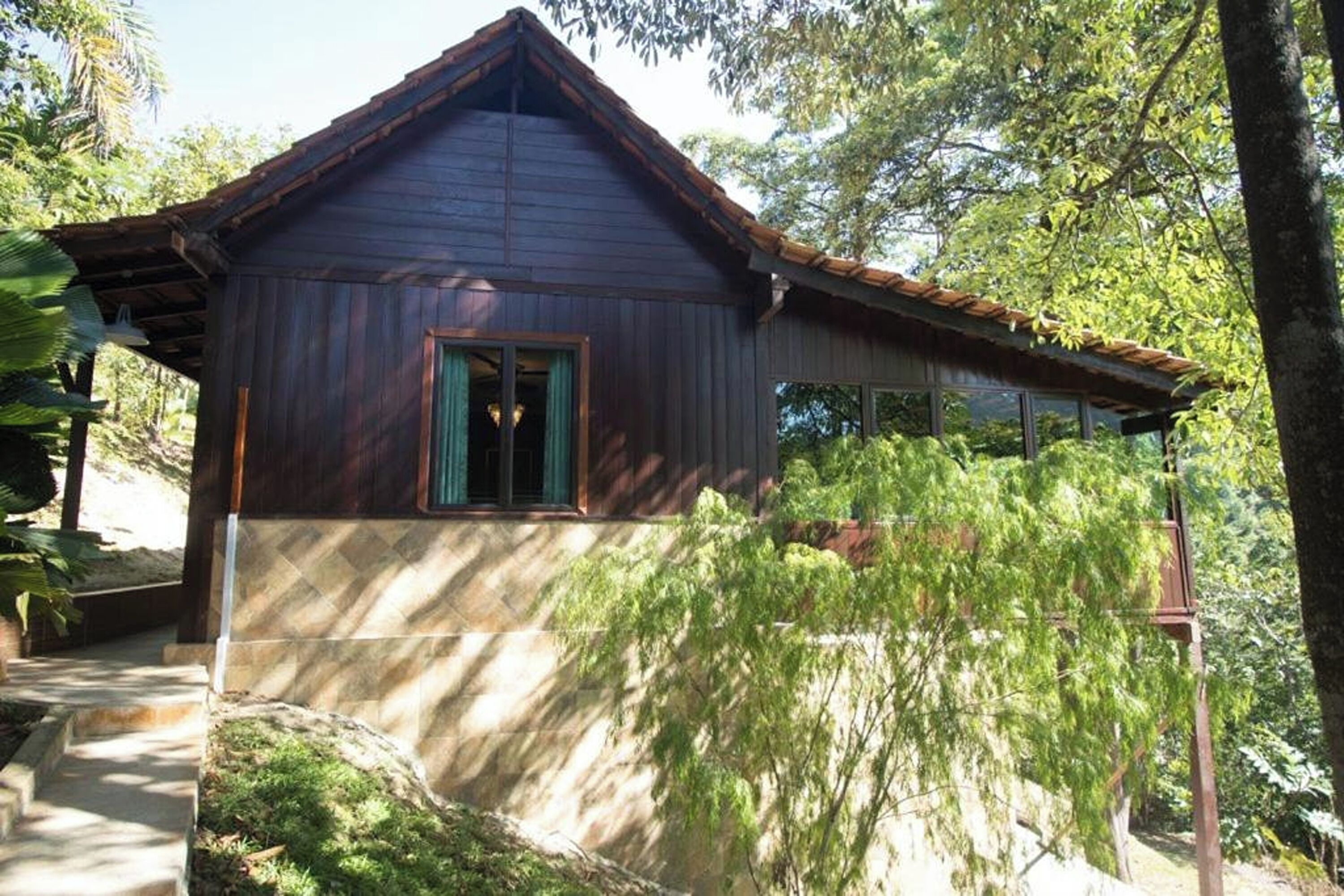 A wooden cabin with a pitched roof, a visible window with green curtains, and a stone foundation.