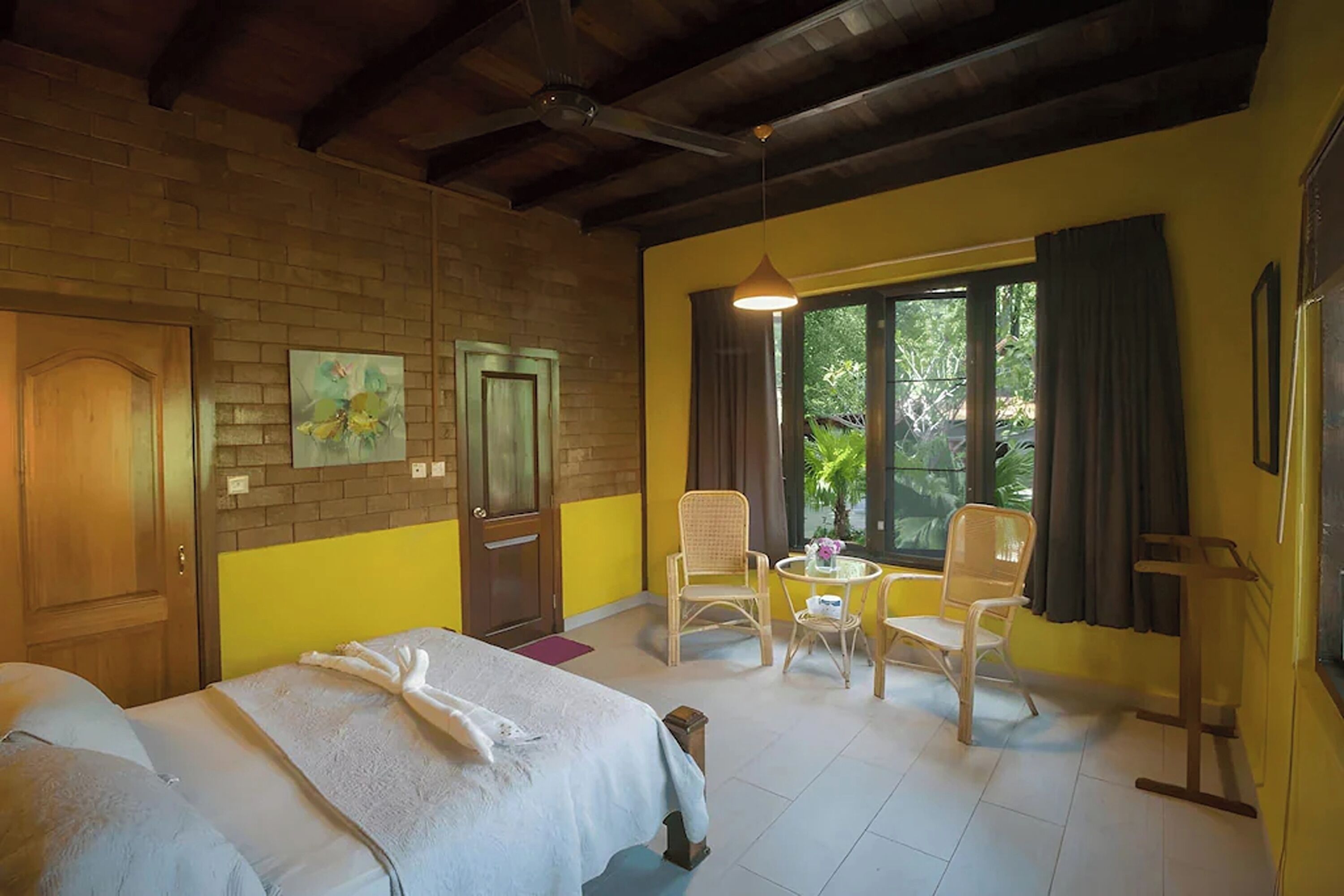 Une chambre d’hôtel avec un lit, deux chaises, une petite table, une fenêtre avec des rideaux et un ventilateur de plafond.