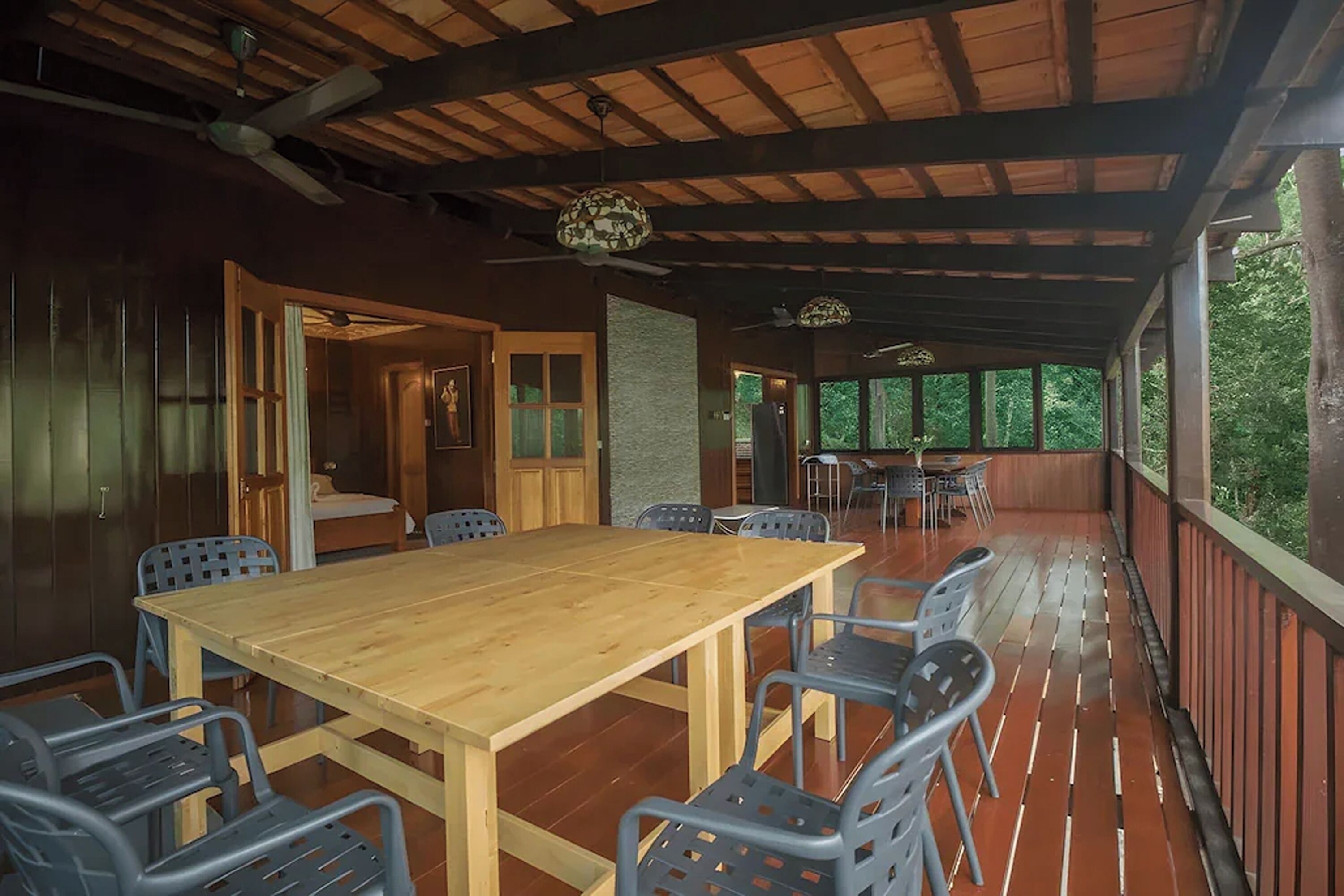 Un espacio de comedor al aire libre con techo, una mesa y sillas de madera, un ventilador de techo y vistas al entorno verde.