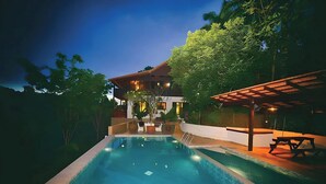 Ein Pool mit überdachtem Bereich, ein Gebäude mit rotem Dach und umgebende Bepflanzung.