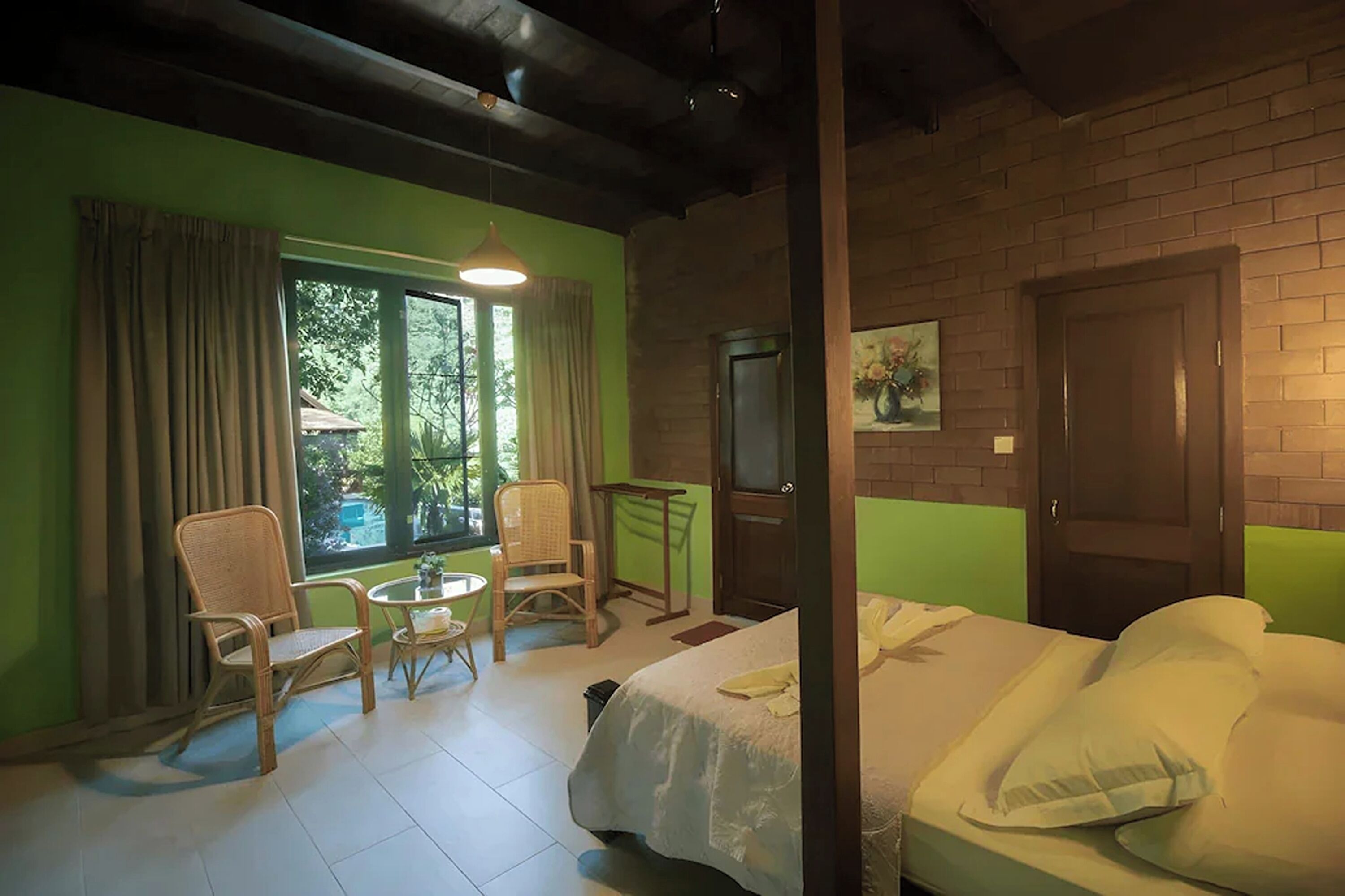 A room with a bed, two chairs, a small table, a window with curtains, a door, and a painting on the wall.