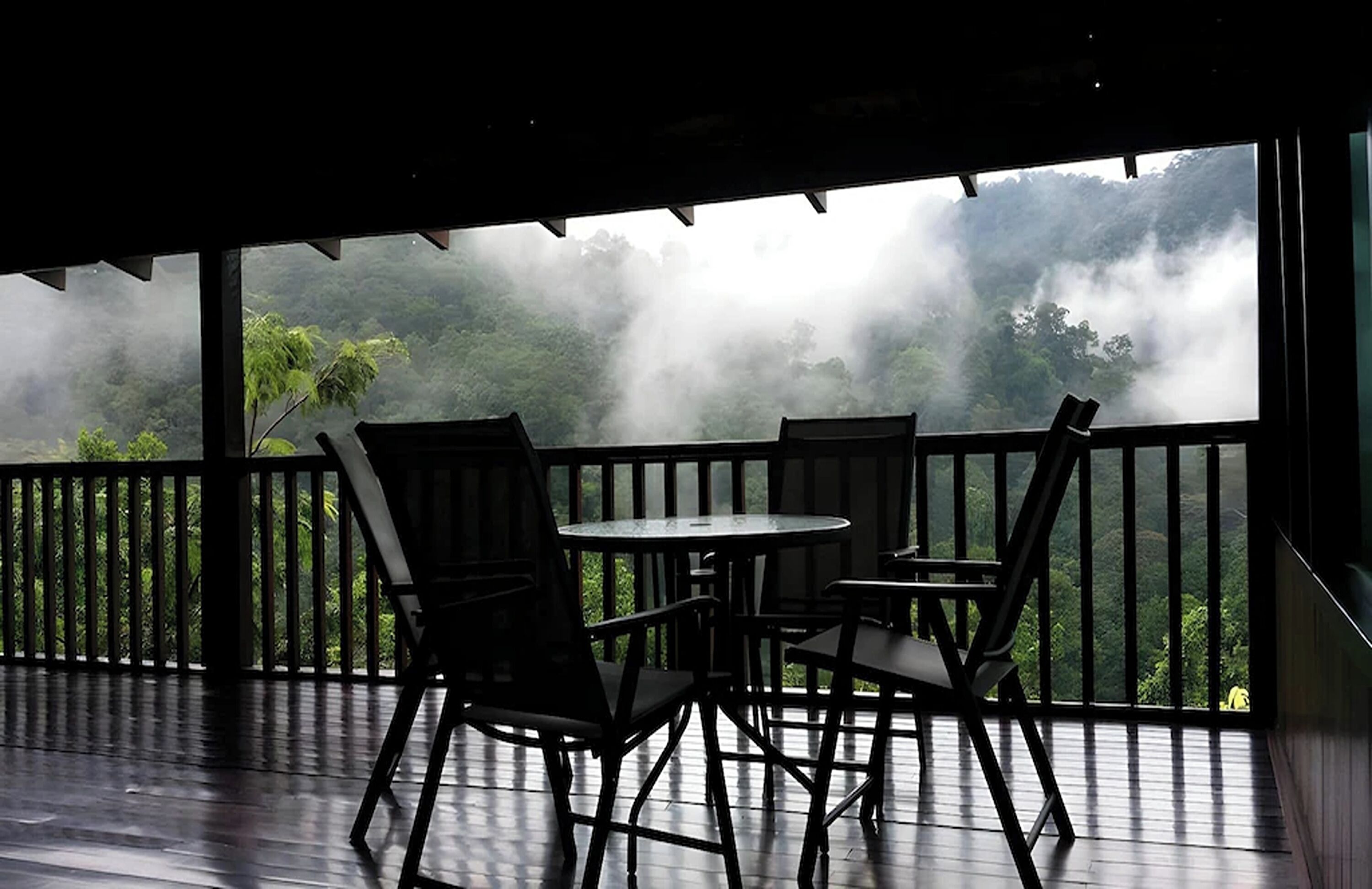 Un espacio de estar al aire libre cubierto, con una mesa y sillas, con vistas a un paisaje boscoso cubierto de niebla.