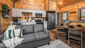 A cozy cabin interior with a wooden interior, a kitchenette, a gray sofa, and a dining table set for four. - New - Dollywood - Fire pit - Arcade - Family Getaway (Sevierville)