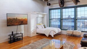 A modern bedroom with a bed, a TV, a white wardrobe, a glass coffee table, and large windows offering a view of the city. - Luxury Loft in the Rochester Public Market! (Rochester)