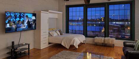 Un soggiorno moderno con un letto, una TV, un tavolino in vetro e vista sulla città attraverso ampie finestre.