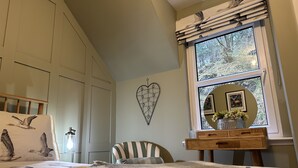 Een slaapkamer met een bed, een stoel, een dressoir, een raam met uitzicht op bomen, en een hartvormige decoratie aan de muur.