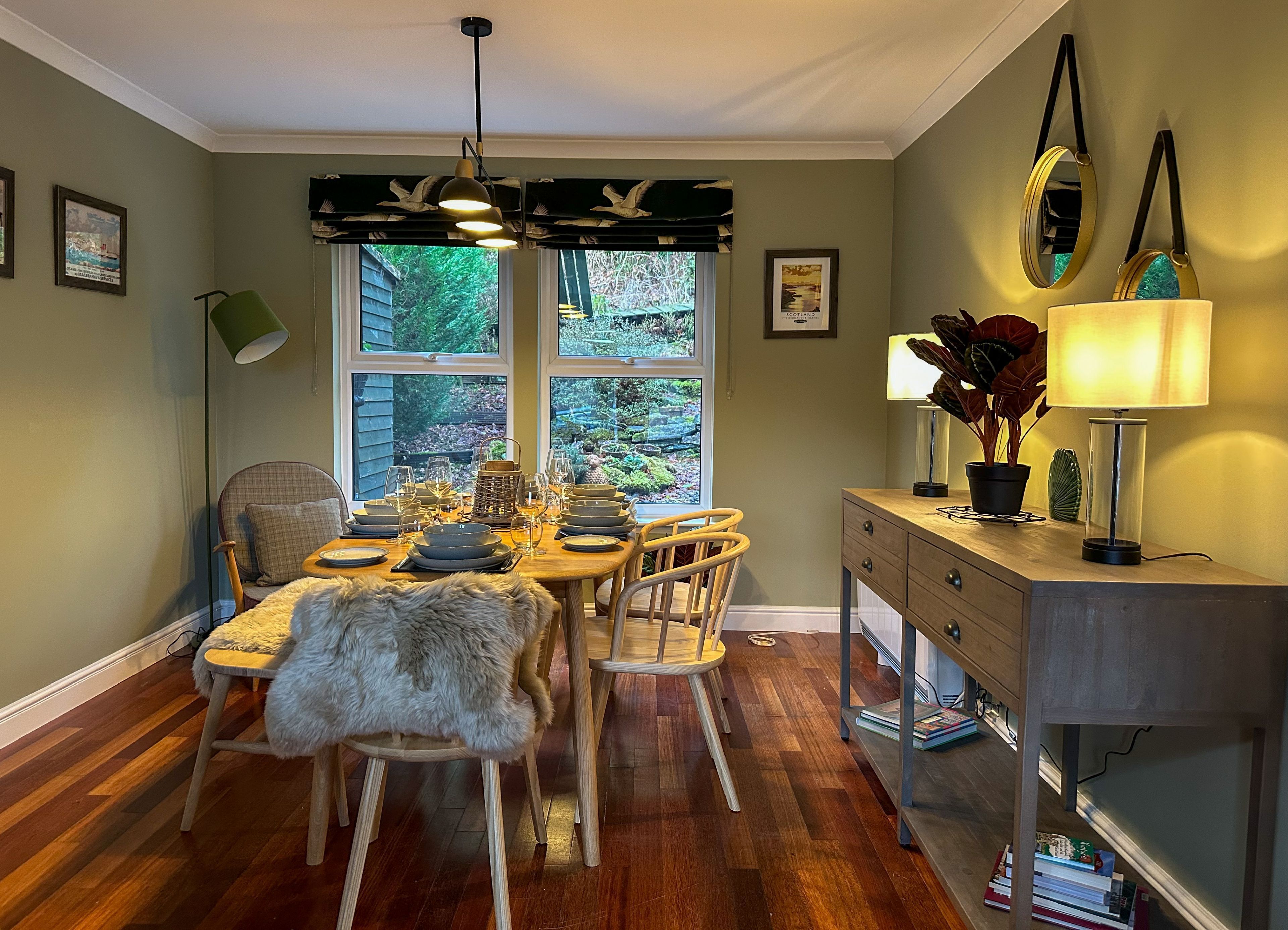 A dining area with a wooden table set for four, surrounded by chairs. There is a window with a view of greenery outside.